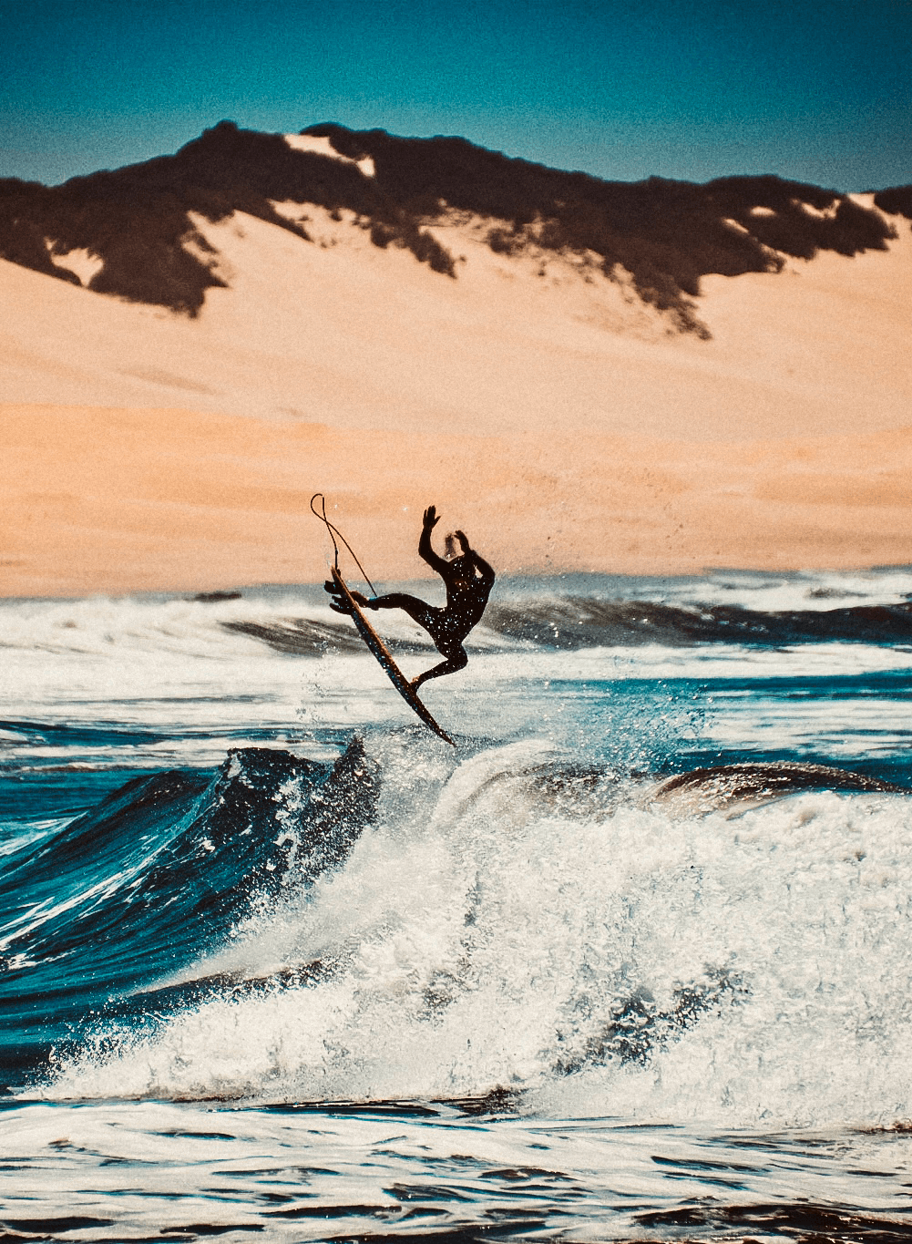 A surfer catching air above a wave, dunes in the background