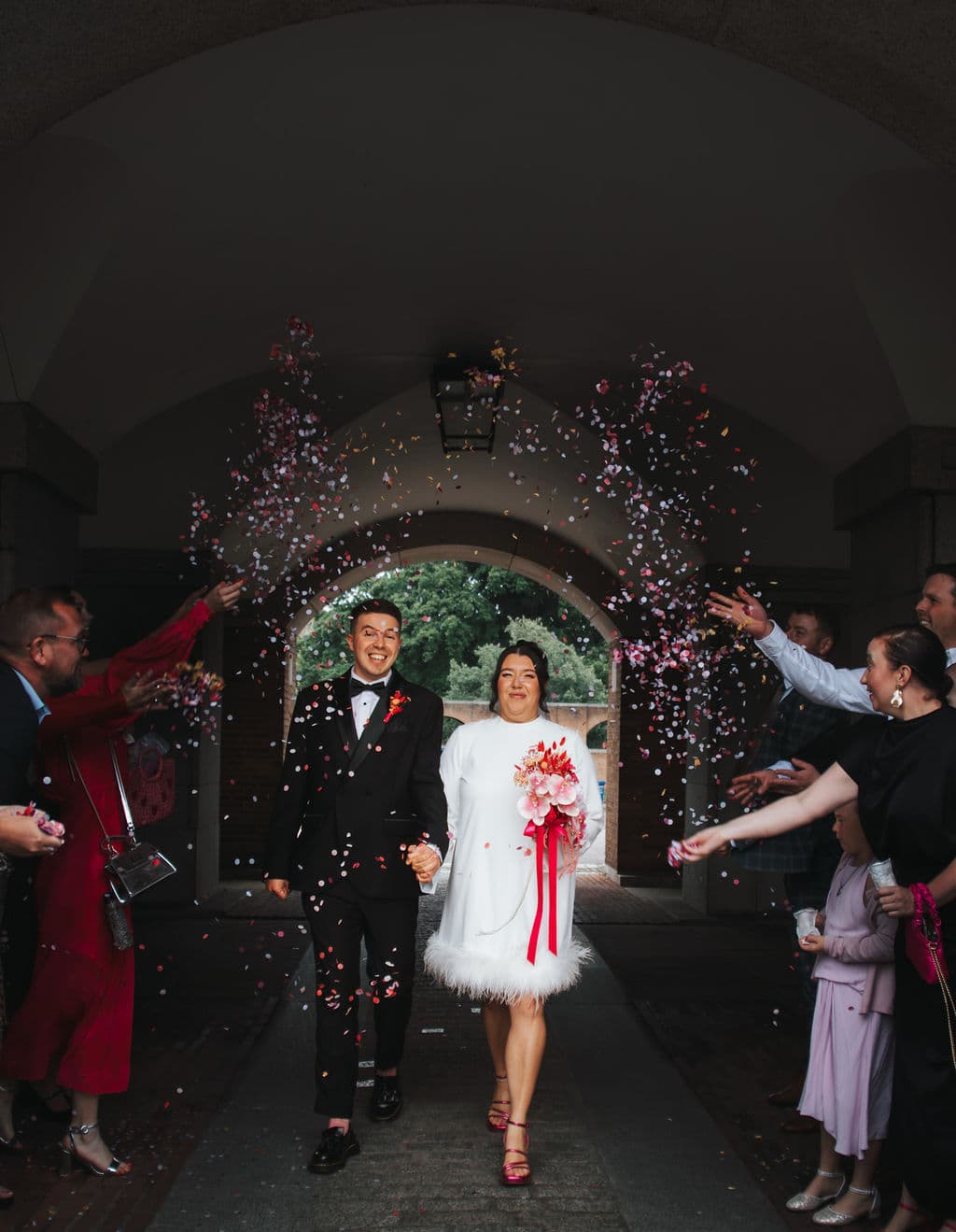 A newly married couple walking through a shower of confetti under a stone archway