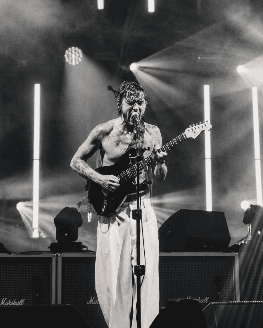A shirtless tattooed musician singing and playing guitar on stage