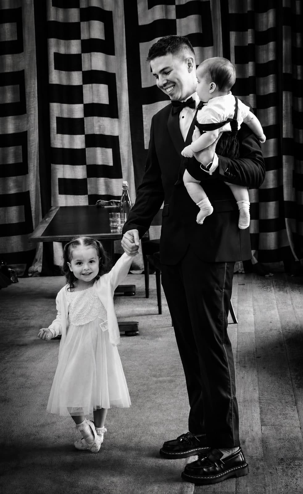 A man in a tuxedo with a baby in one arm, a small girl in ballet shoes spinning in the other hand