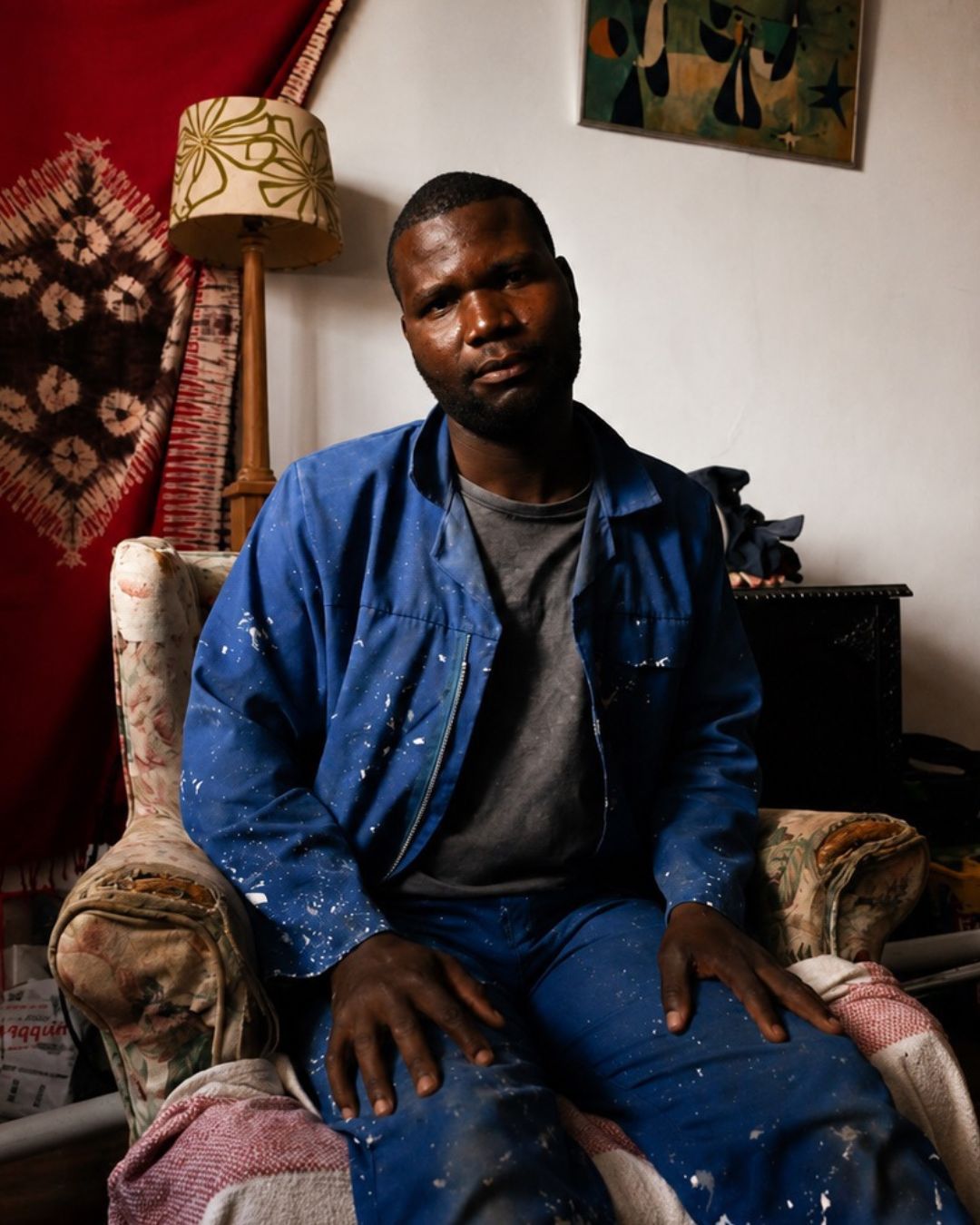 Man in blue paint-splattered overalls seated in armchair, documentary portrait, South Africa