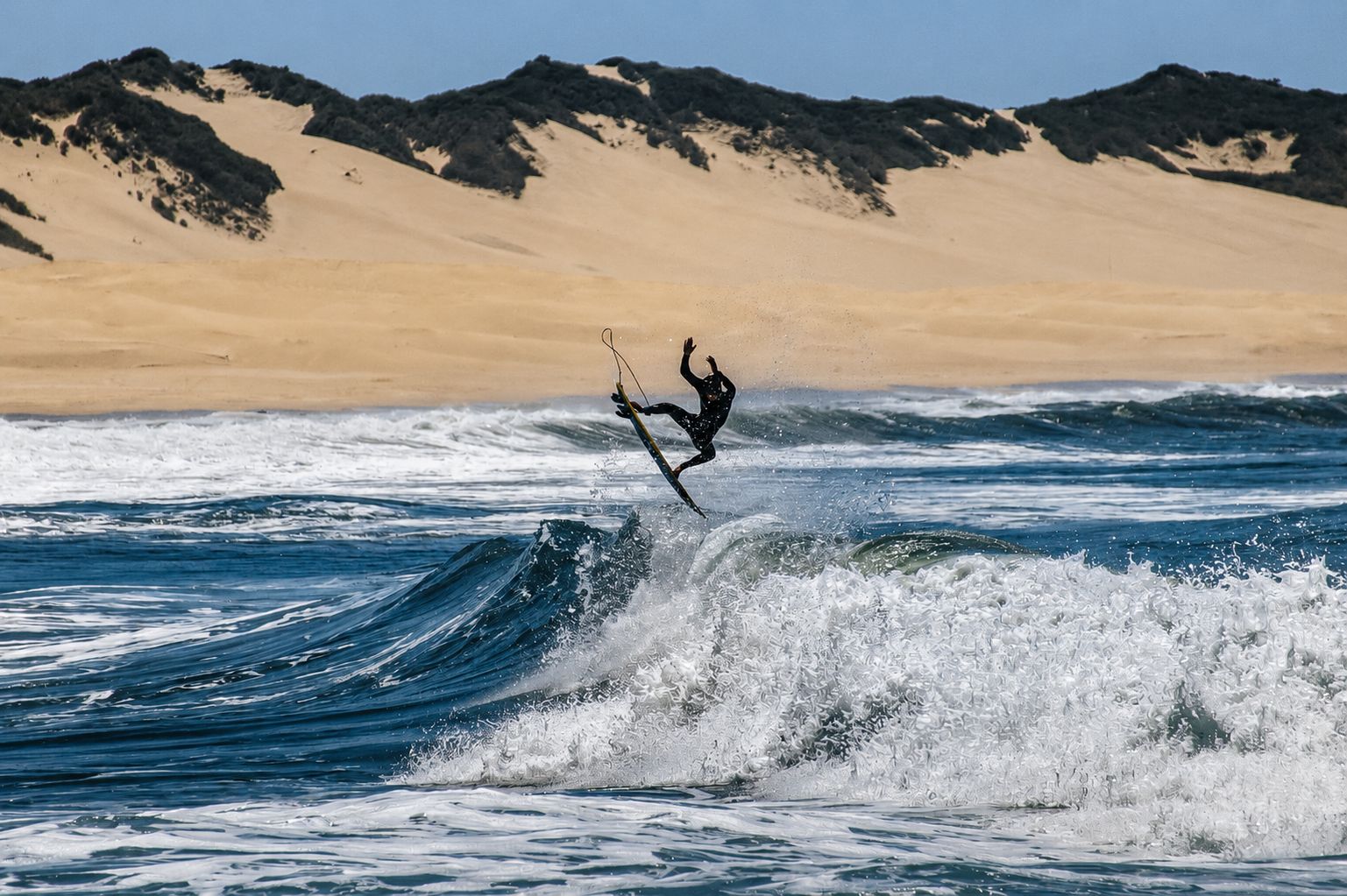 Surfer launching air above wave, Eastern Cape dunes behind