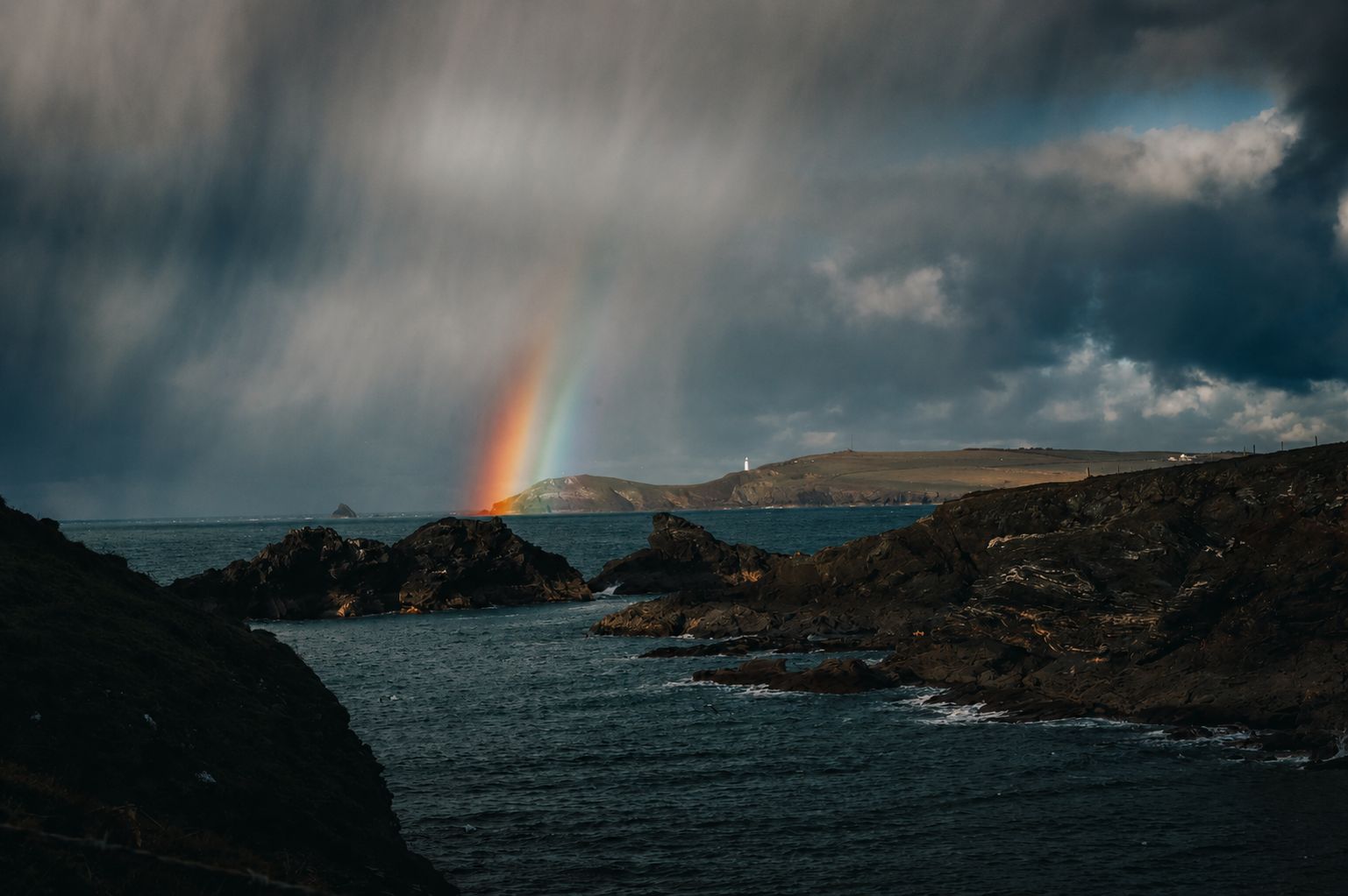 Cornwall coastline with dramatic rainbow over dark storm clouds and rocky coves